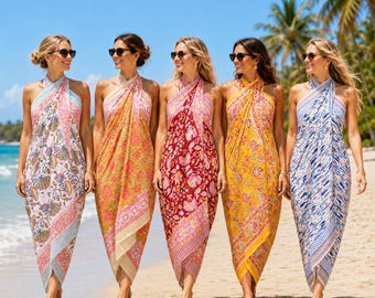 May include: Five sleeveless halter-neck dresses featuring vibrant floral and geometric patterns in shades of pink, orange, red, yellow, and blue. Each dress has a wrap-style skirt with a side slit, worn with dark sunglasses on a sandy beach.