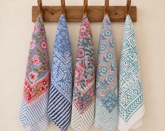 May include: Five rectangular cotton kitchen towels with diverse block-printed patterns in shades of blue, pink, green, and grey, hanging from a wooden wall-mounted rack with five hooks. Each towel features unique floral, geometric, and striped designs.