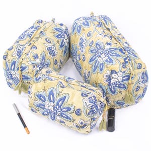 May include: Three quilted cosmetic bags in varying sizes. The bags are yellow with a blue floral pattern. A black eyeliner pencil and a black lipstick tube are next to the bags. Each bag has a tassel.