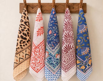May include: Five rectangular fabric napkins hang from a wooden wall rack. The napkins feature diverse patterns including a tan leopard print, a white and orange floral design, a blue and teal botanical print, a white and maroon floral motif, and a blue floral pattern with yellow accents.