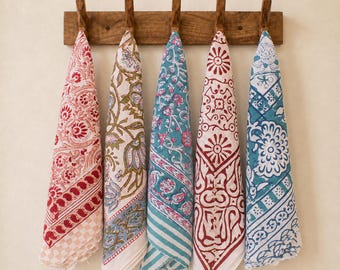 May include: Five rectangular fabric cloths with diverse block-printed patterns in red, blue, green, and white, hanging from a wooden wall rack with five pegs. Each cloth features distinct floral, geometric, and paisley motifs on a light background.