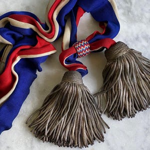 May include: A decorative tieback featuring a long, woven fabric cord in vibrant blue, red, and beige stripes. The cord is secured by a small, multi-coloured chevron-patterned ring and terminates in two large, voluminous, metallic gold-toned tassels.