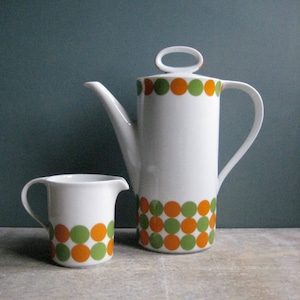 Può includere: Un set da tè in ceramica bianca con un motivo geometrico di cerchi arancioni e verdi. La teiera ha un coperchio con un manico ad anello.