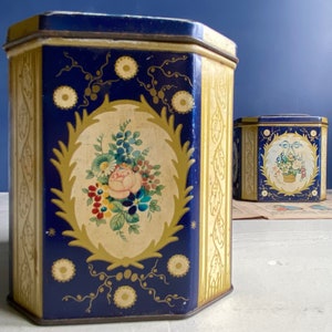 Può includere: Due scatole di latta esagonali vintage con disegni floreali blu e dorati. La scatola più grande presenta un bouquet di fiori con una rosa, mentre la scatola più piccola ha un arrangiamento floreale in un cestino.