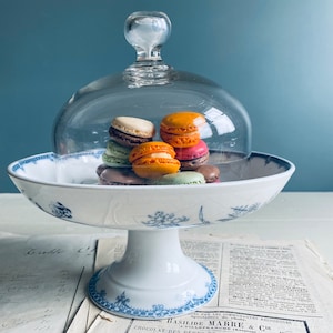 Op de afbeelding: Een witte taartstandaard met een blauw bloemenpatroon en een glazen stolp. De standaard heeft een ronde basis en een hoge, cilindrische voet. De stolp is van helder glas en heeft een knop bovenop. Er zijn kleurrijke macarons gestapeld in de stolp.