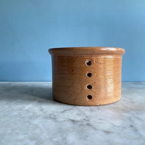 May include: A brown ceramic butter crock with five holes in the side. The crock is sitting on a white marble surface.