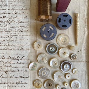 Op de afbeelding: Een verzameling naaibenodigdheden waaronder een houten klosje met bruin garen, een donkerpaarse houten stempel met de letters MH, twee ronde garenklosjes, een lange botkleurige naald en diverse witte en doorschijnende parelmoeren knopen.