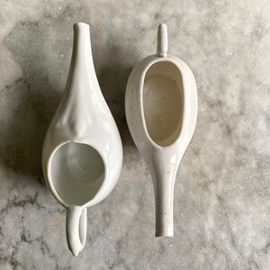 May include: Two white ceramic enema bulbs, one with a rounded spout and the other with a more pointed spout.