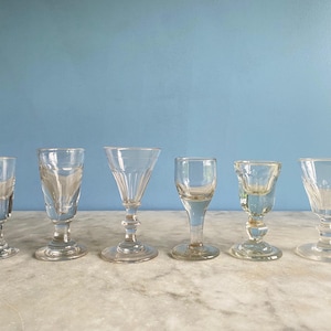 May include: Six clear glass cordial glasses of varying shapes and sizes are arranged in a row. The glasses have stems and decorative detailing. The background is a solid light blue.