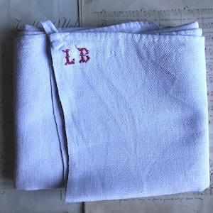 Può includere: Un asciugamano da cucina in lino bianco con un motivo a spina di pesce e le iniziali "LB" ricamate in filo rosso.