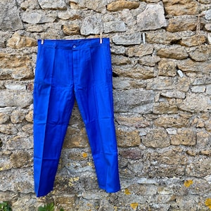 Può includere: Pantaloni blu brillante appesi a un filo contro un muro di pietra. I pantaloni sono di colore pieno, con chiusura a bottone e tasche. I pantaloni sono realizzati in un tessuto liscio e sembrano nuovi. I pantaloni sono di un colore vivace e accattivante.