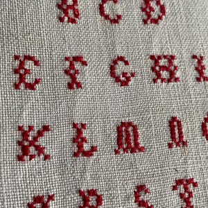 May include: A close-up of a white fabric with red embroidered letters of the alphabet. The letters are stitched in a simple cross-stitch pattern.