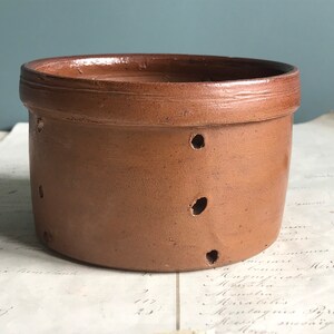Peut inclure: Un pot en terre cuite brun avec des trous sur les côtés. Le pot est posé sur une surface blanche avec de l'écriture dessus.