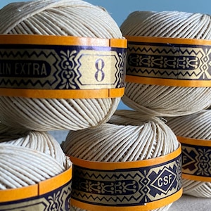 Op de afbeelding: Vier bollen gebroken wit linnen garen, elk omwikkeld met een decoratieve oranje en donkerblauwe papieren band. De banden bevatten de tekst FIL DE LIN EXTRA, het cijfer 8 en de letters CSF, samen met ingewikkelde geometrische patronen.