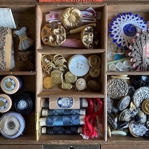 Pode incluir: Um organizador de madeira cheio de artigos de costura. Os compartimentos contêm carretéis de linha, botões, enfeites de metal vintage e outros materiais de artesanato. A paleta de cores inclui dourado, azul, branco e preto.