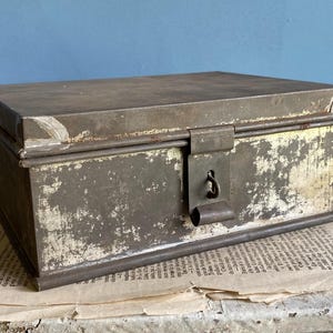 Może przedstawiać: Antyczne metalowe pudełko z pokrywką na zawiasach i zamkiem. Pudełko jest pomalowane na wyblakły szary kolor, z prześwitującą białą farbą. Pudełko ma zużyty, rustykalny wygląd.