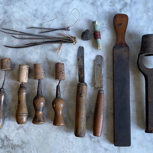 May include: A collection of vintage leatherworking tools including a bundle of metal needles, a small spool of red thread, four awls with wooden handles and cork tips, two knives with wooden handles, a long wooden strop, and a heavy metal punch tool.