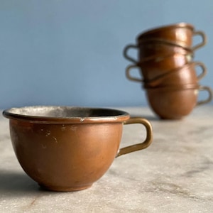 May include: A set of four small copper cups with brass handles. The cups are stacked on top of each other and have a worn, vintage look.