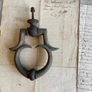 May include: A weathered bronze door knocker featuring a circular ring design with a central mounting post and a threaded nut at the top. The metal surface displays a dark, aged patina with visible signs of oxidation and wear throughout the piece.