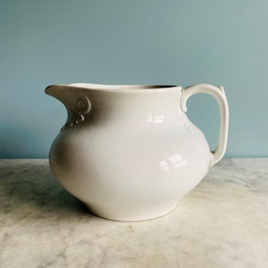 May include: A white ceramic pitcher with a rounded body, a curved handle, and a prominent pouring spout. The exterior surface features subtle embossed scrollwork details near the rim and handle attachment point, with a smooth finish.