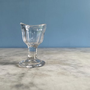 Könnte beinhalten: Ein klarer Augenbad aus Glas mit ausgestelltem Rand und kleinem, rundem Sockel. Das Vintage-Augenbad hat vertikale Rillen auf der Schale. Es steht auf einer Marmoroberfläche mit blauem Hintergrund.