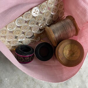 May include: A pink fabric pouch containing sewing essentials. The pouch holds a strip of white buttons, two spools of thread, and two spools of black thread. One black thread spool has the text "120 Extra S".
