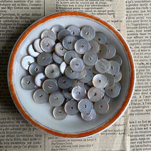 Peut inclure: Gros plan d'une assiette en céramique blanche remplie d'une collection de boutons ronds en nacre gris. Les boutons ont un trou au centre et sont disposés de manière aléatoire.