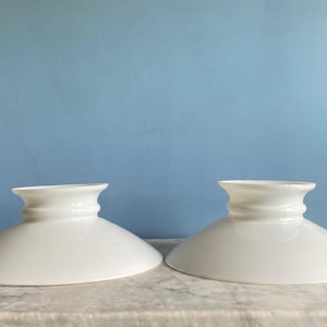 Op de afbeelding: Twee witte glazen lampenkappen met een uitlopende, komachtige vorm. Elke kap heeft een decoratieve, geribbelde hals en een brede, naar beneden hellende rand. De kappen worden gepresenteerd op een marmeren oppervlak tegen een blauwe achtergrond.
