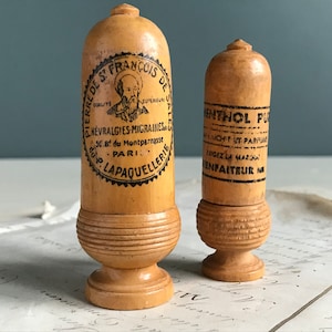 May include: Two vintage wooden medicine containers with labels. The larger container is labeled "Pierre de S. Francois de Sales, Qualite Superieure, Nevrgalgies-Migraines, 50. Bd du Montparnasse, Paris, P. Lapaquellerie". The smaller container is labeled "Menthol Pur, Sachet et Parfume, Exigez la Marque, Infaiteur Par".