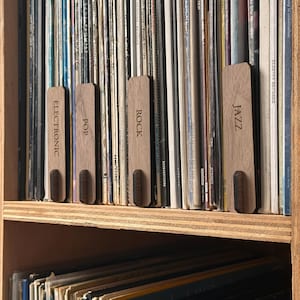 May include: A wooden shelf with vinyl records organised by genre. The genres are labelled with wooden tags: Jazz, Rock, Pop, and Electronic.