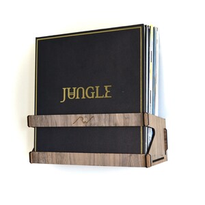 May include: A wooden wall-mounted record shelf with a stack of vinyl records. The top record has the word "JUNGLE" printed in gold lettering.