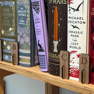 May include: A selection of books held in place by wooden bookends labelled A, B, C, and D. The books have colourful covers and titles including "Jane Austen" and "Jurassic Park".