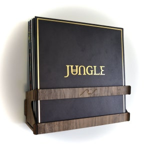 Puede incluir: Un estante de discos de madera marrón oscuro con un borde dorado, que contiene una pila de discos de vinilo. El disco superior tiene una inscripción dorada que dice "JUNGLE".