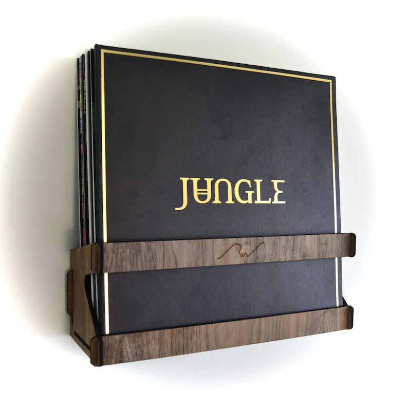 May include: A dark brown wooden record shelf with a gold trim, holding a stack of vinyl records. The top record has a gold inscription that reads "JUNGLE".