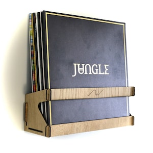 May include: A wooden wall-mounted record shelf with two tiers, holding vinyl records. The top record has a black cover with gold lettering that reads "JUNGLE".
