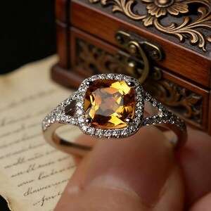 Pu&ograve; includere: Un anello d'argento con una grande pietra preziosa quadrata color ambra al centro, circondata da un alone di pietre pi&ugrave; piccole e trasparenti. La fascia dell'anello presenta ulteriori piccole pietre. L'anello &egrave; tenuto davanti a un cofanetto di legno.