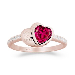 Può includere: Anello in oro rosa con una pietra preziosa rossa a forma di cuore. L'anello presenta l'iscrizione "Love You MOM" ed è impreziosito da piccole pietre preziose trasparenti lungo la fascia. L'anello è posto su uno sfondo bianco.