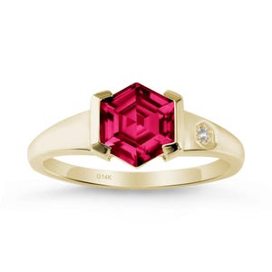 May include: A gold ring with a striking, hexagonal, deep red gemstone at its centre. A small, clear, round stone is set on the side. The band is polished gold. The inscription "G14K" is visible on the band.