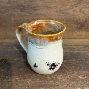 May include: A ceramic mug with a rounded body and a handle, featuring a cream-colored base with a speckled texture. The upper rim is glazed in a warm, drippy amber and brown tone. A small black bee illustration is printed on the front surface.