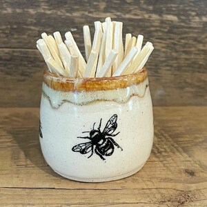 May include: A small, round, cream-colored ceramic holder featuring a black bee illustration on the front. The rim has a decorative brown drip glaze finish. The container is filled with a cluster of light-colored wooden toothpicks standing upright.