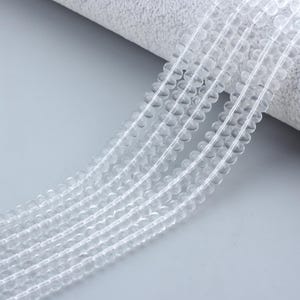 May include: Strands of clear, round beads. The beads are strung together, creating multiple lines of the same beads. The beads are transparent and appear to be made of glass or crystal. The background is a light gray color.