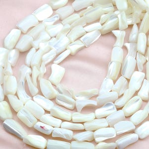 Puede incluir: Primer plano de un collar hecho de cuentas rectangulares blancas de nácar. Las cuentas tienen un brillo iridiscente y están ensartadas para formar un collar. El fondo es de tela rosa suave.