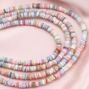May include: Strands of pastel-colored shell beads in shades of pink, blue, green, and tan. The beads are flat and disc-shaped, strung together to form a necklace or bracelet. The background is a soft pink fabric.