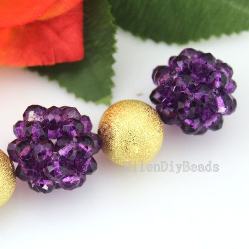 May include: Close-up of beads in a variety of shapes and colors. Two faceted, deep purple beads are next to a gold, textured bead. The beads are on a white surface with a blurred background of a red flower and green leaves.