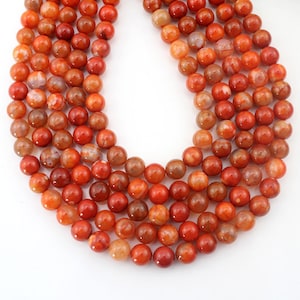 Puede incluir: Una hebra de cuentas redondas de ágata naranja con vetas blancas y marrones. Las cuentas están enhebradas en un hilo transparente y tienen un diámetro aproximado de 8 mm.
