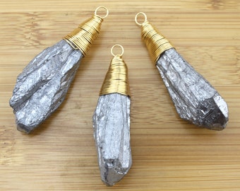 May include: Three raw, silver-colored crystal pendants with gold wire wrapping. Each pendant has a loop at the top for hanging. The crystals have a rough, natural texture and are displayed on a wooden surface.