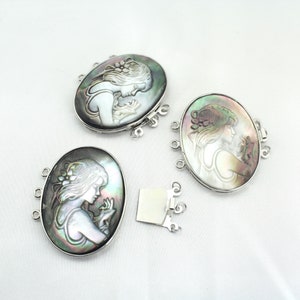 Puede incluir: Tres cierres ovalados de plata con incrustaciones de nácar que representan una imagen tallada de una mujer. Cada cierre tiene tres lazos de plata para sujetar a un collar o pulsera.
