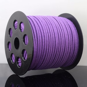 May include: A spool of purple suede cord. The cord is tightly wound around a black plastic spool. The cord is a vibrant shade of purple, and the texture appears soft and velvety. The spool is sitting on a reflective surface.