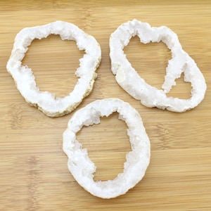 May include: Three white crystal agate slices with a hole in the center. The slices are on a light brown wooden surface.