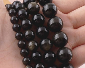 Könnte beinhalten: Eine Ansammlung glatter, kugelförmiger schwarzer Obsidianperlen mit subtilen goldenen Glanzreflexen. Die Perlen variieren in der Größe und sind in einer Gruppe angeordnet, was eine polierte, reflektierende Oberfläche mit natürlichen dunklen Tönen zeigt.